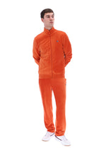 A man modeling a matching orange corduroy tracksuit, consisting of a zip-up jacket and matching pants, paired with white sneakers, against a plain white background.