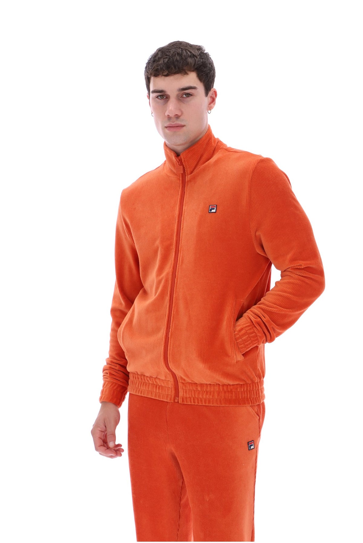 A man modeling a bright orange corduroy tracksuit, featuring a zip-up jacket and matching pants, against a plain white background.