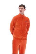 A man wearing a bright orange velour tracksuit, featuring a zip-up jacket with a small embroidered logo on the chest and matching pants, shot against a solid white background.