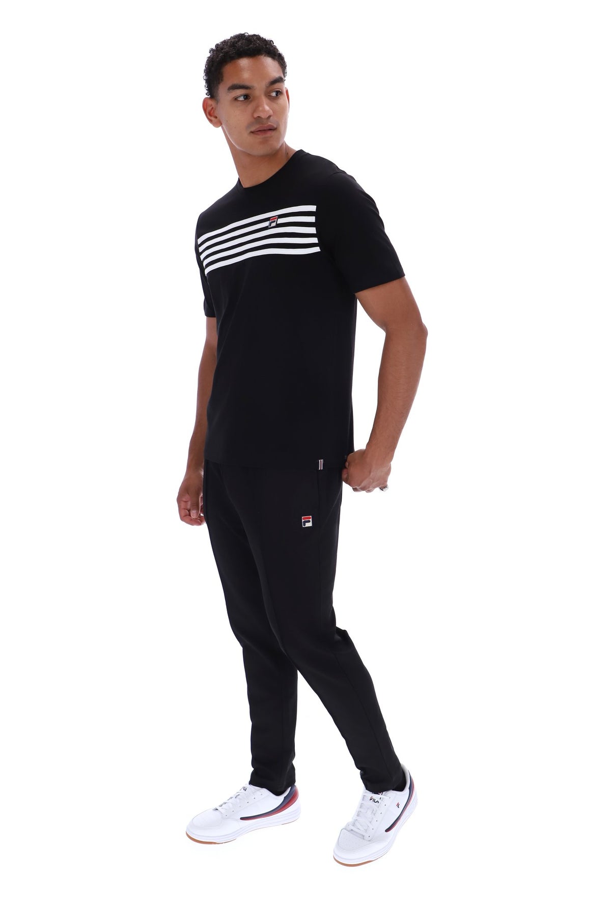 A man modeling a black short-sleeve t-shirt with three white horizontal stripes across the chest, paired with black joggers featuring a small red and white logo on the left thigh, and white sneakers, set against a plain white background.