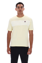 A man wearing a pale yellow short-sleeve t-shirt with white horizontal stripes across the chest and a small red, white, and blue Fila logo on the left chest, paired with black pants, against a plain white background.