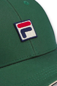 Close-up of a green baseball cap featuring a square embroidered patch with the Fila logo in red, white, and navy blue.