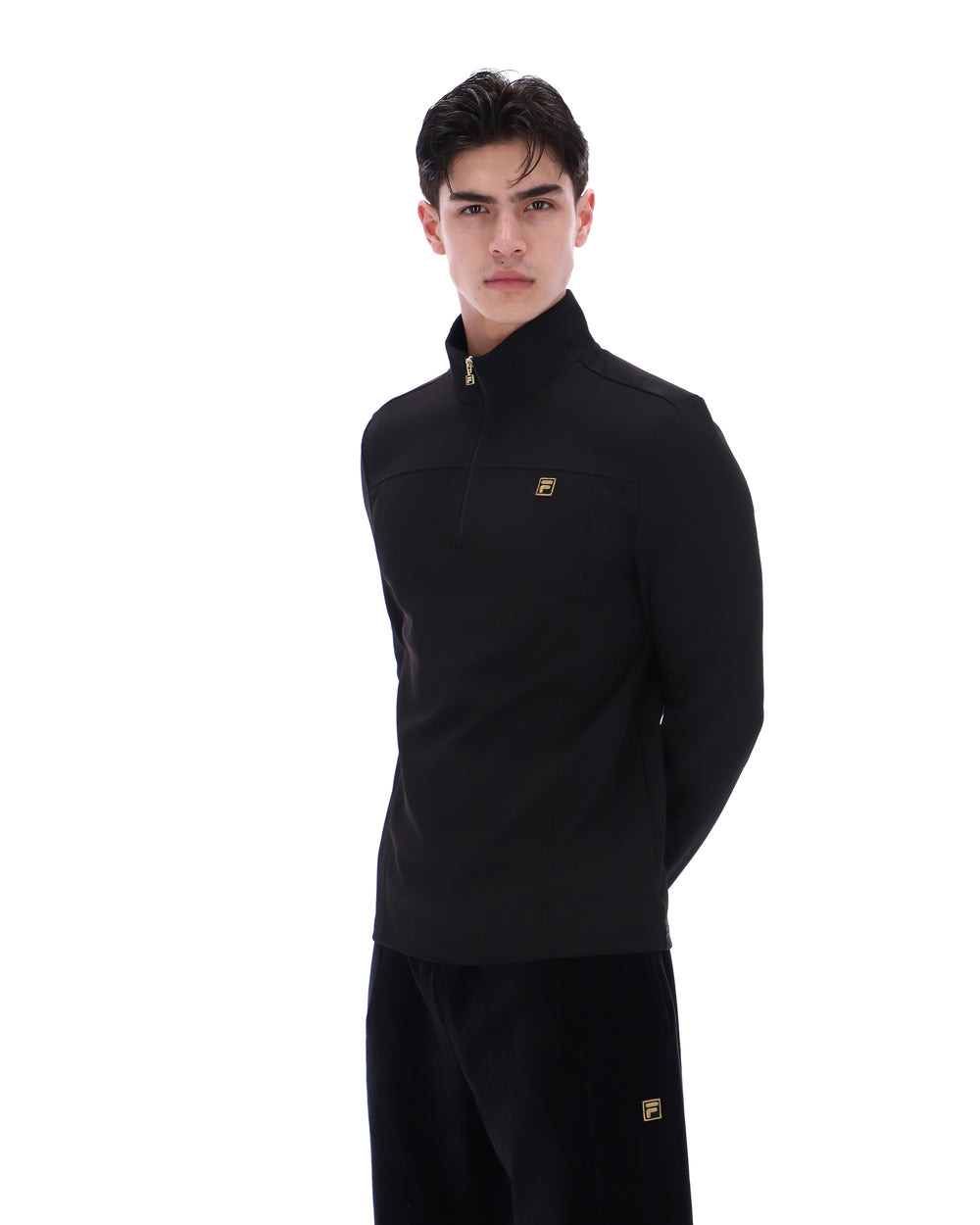 Man wearing a black jacket with a FILA gold logo, paired with matching bottoms, against a white background