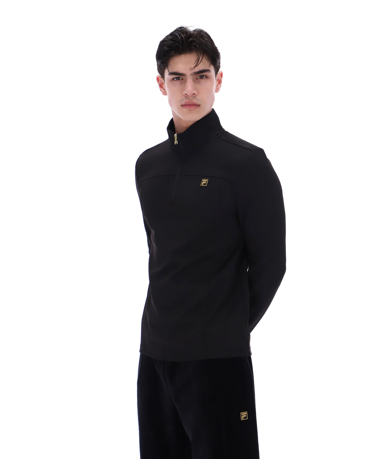 Man wearing a black jacket with a FILA gold logo, paired with matching bottoms, against a white background