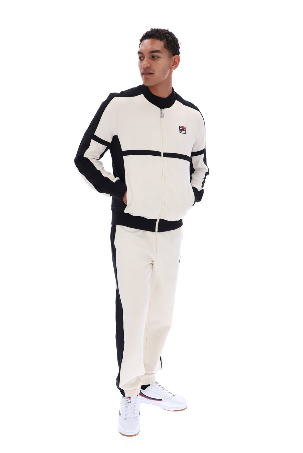 A man modeling a matching cream and black Fila tracksuit, featuring a zip-up jacket with a small Fila logo on the chest and matching jogger pants, set against a plain white background.