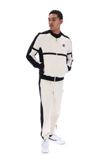 A man modeling a matching cream and black Fila tracksuit, featuring a zip-up jacket with a small Fila logo on the chest and matching jogger pants, set against a plain white background.