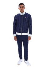 A man modeling a navy blue pinstriped zip-up jacket with white ribbed trim, matching navy pants, and white sneakers against a plain white background.