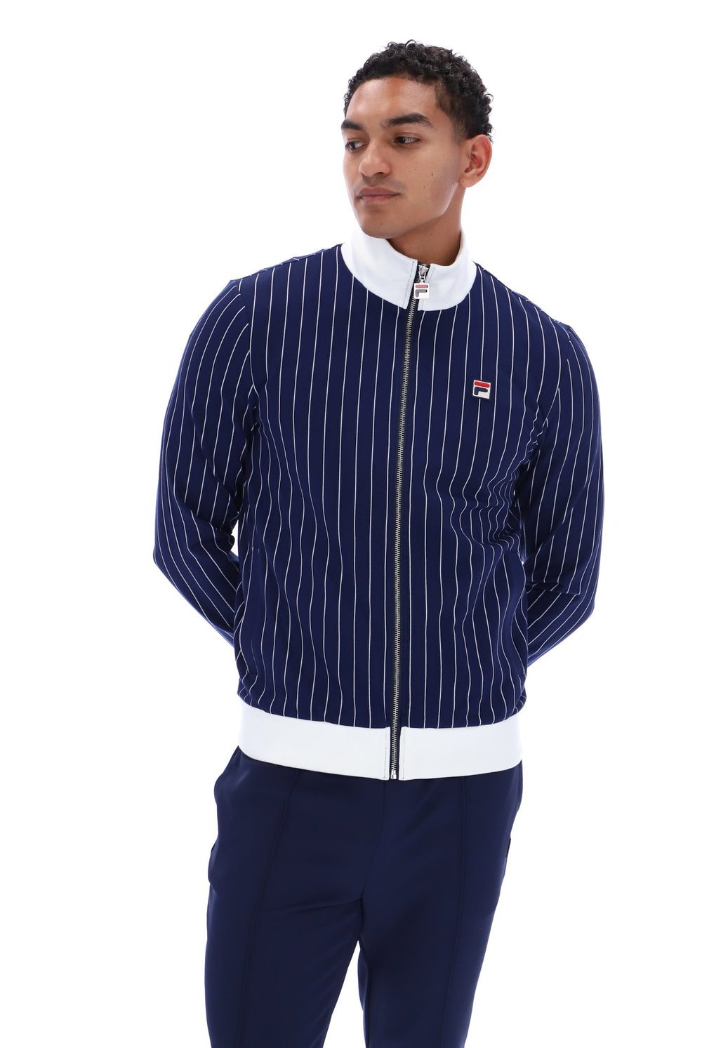 A man wearing a navy blue pinstriped zip-up jacket with white ribbed trim and a small Fila logo on the chest, paired with navy pants, against a plain white background.