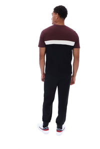 A man viewed from behind wearing a short-sleeve t-shirt with a maroon upper section, a white horizontal stripe across the back, and black pants, paired with white sneakers.