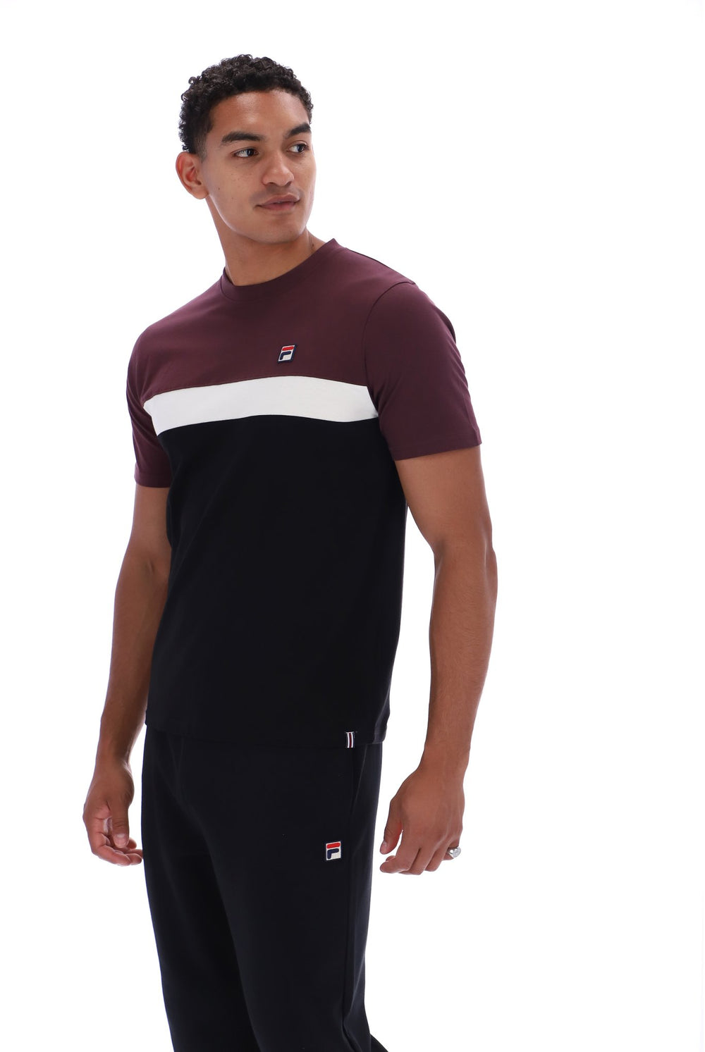 A man wearing a color-blocked short-sleeve t-shirt and matching sweatpants by Fila. The t-shirt features a maroon upper chest and shoulders, a wide white horizontal stripe across the chest, and a solid black lower body.