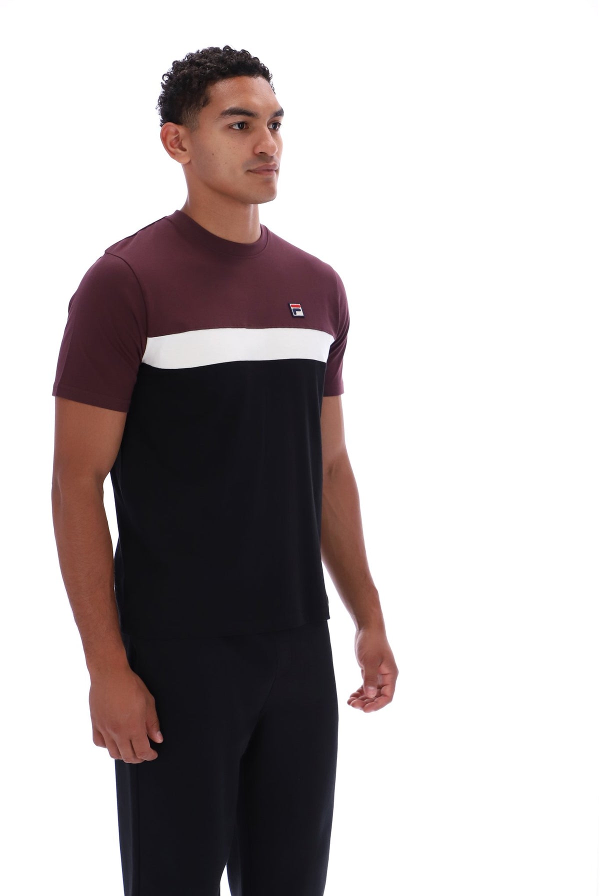 A man wearing a color-blocked short-sleeve t-shirt and matching dark trousers, standing against a plain white studio background. The t-shirt features a maroon upper section, a white horizontal stripe across the chest, and a small red and blue logo on the left chest.