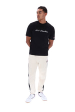 A man modeling a black Champion t-shirt with '1911 Italia' printed on the chest, paired with off-white track pants featuring black side stripes and white sneakers, shot against a solid white background.