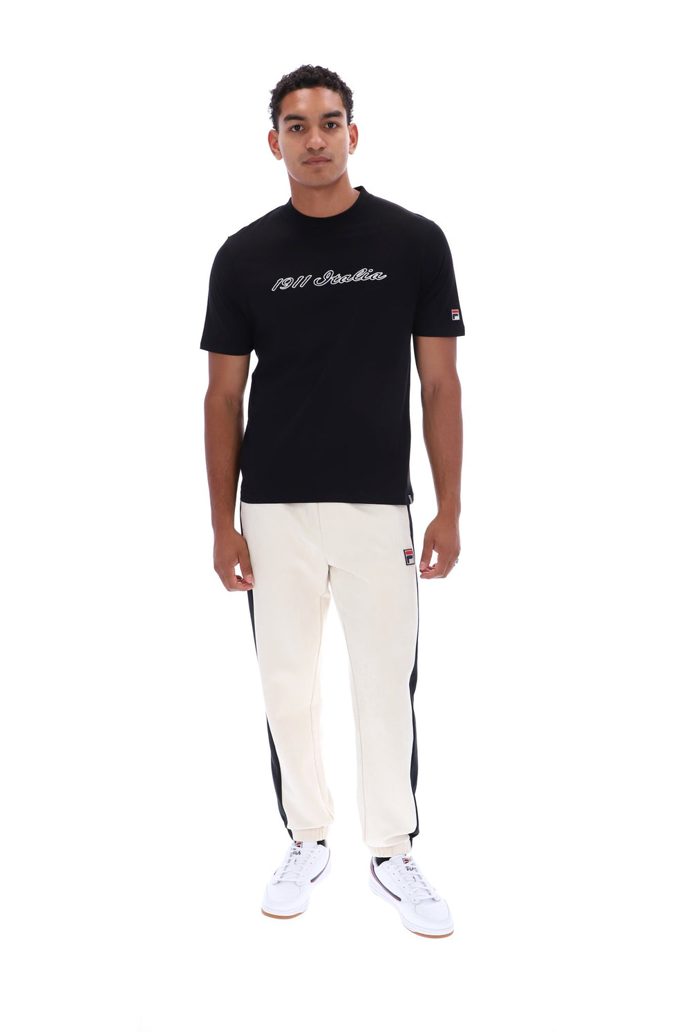 A man modeling a black crewneck t-shirt with white script text on the chest, paired with off-white track pants featuring black side stripes and white sneakers, shot against a solid white background.