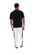 A man viewed from behind wearing a black short-sleeve t-shirt and off-white track pants with black side panels, standing against a plain white background.