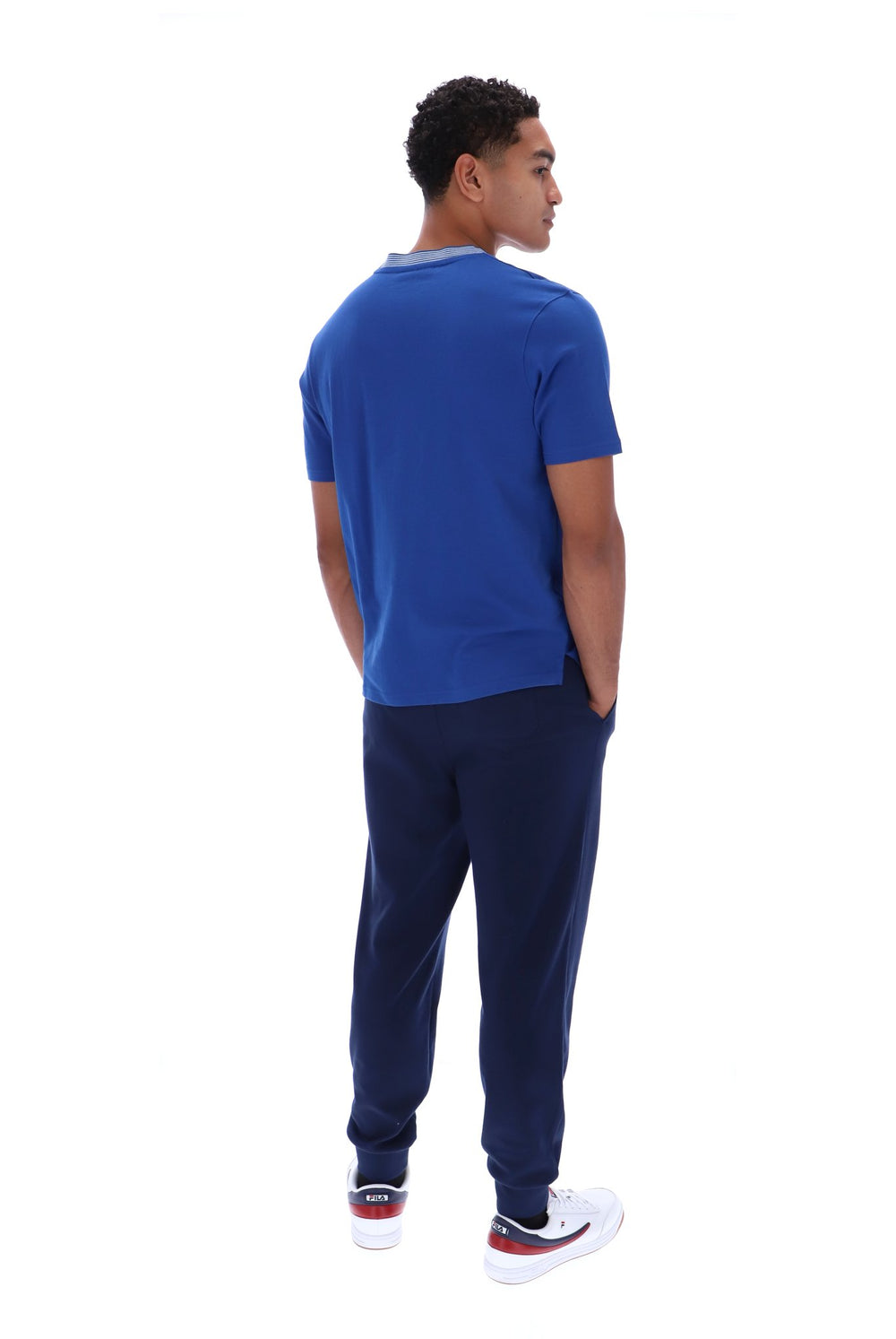 A man shown from the back wearing a royal blue short-sleeve t-shirt, navy blue jogger pants, and white sneakers with red accents, set against a plain white background.