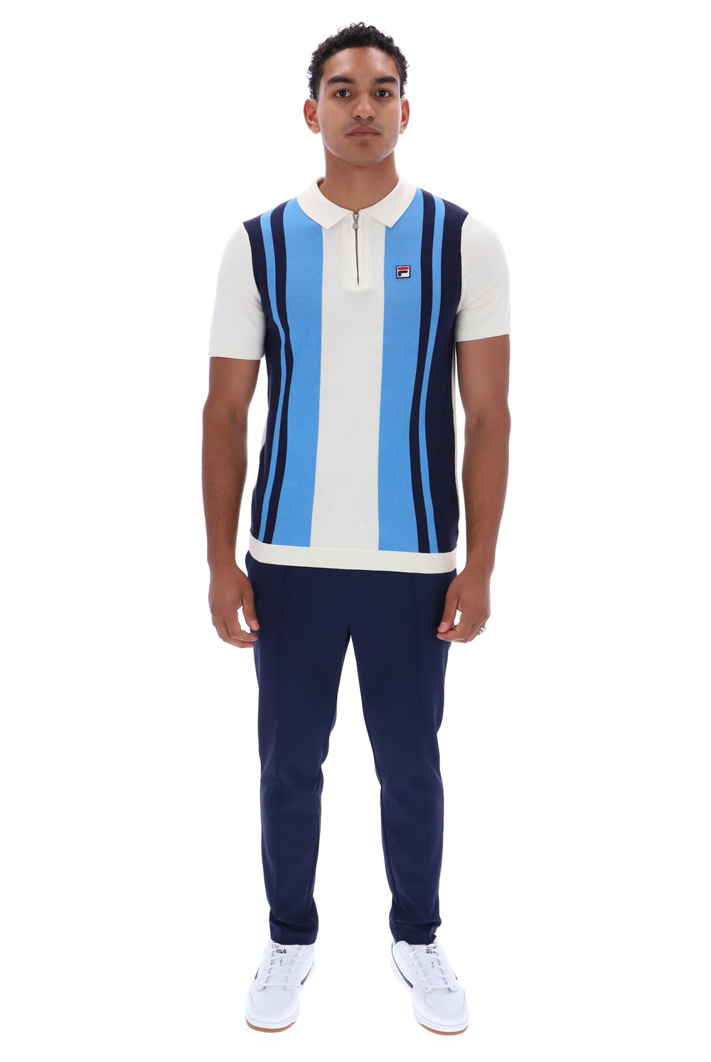 A man modeling a FILA short-sleeve polo knit vest with vertical light blue, white, and navy stripes, paired with navy pants and white sneakers, on a solid white background.