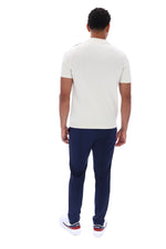 A man viewed from behind wearing a cream short-sleeve polo shirt, navy blue trousers, and white sneakers with red accents, set against a plain white background.