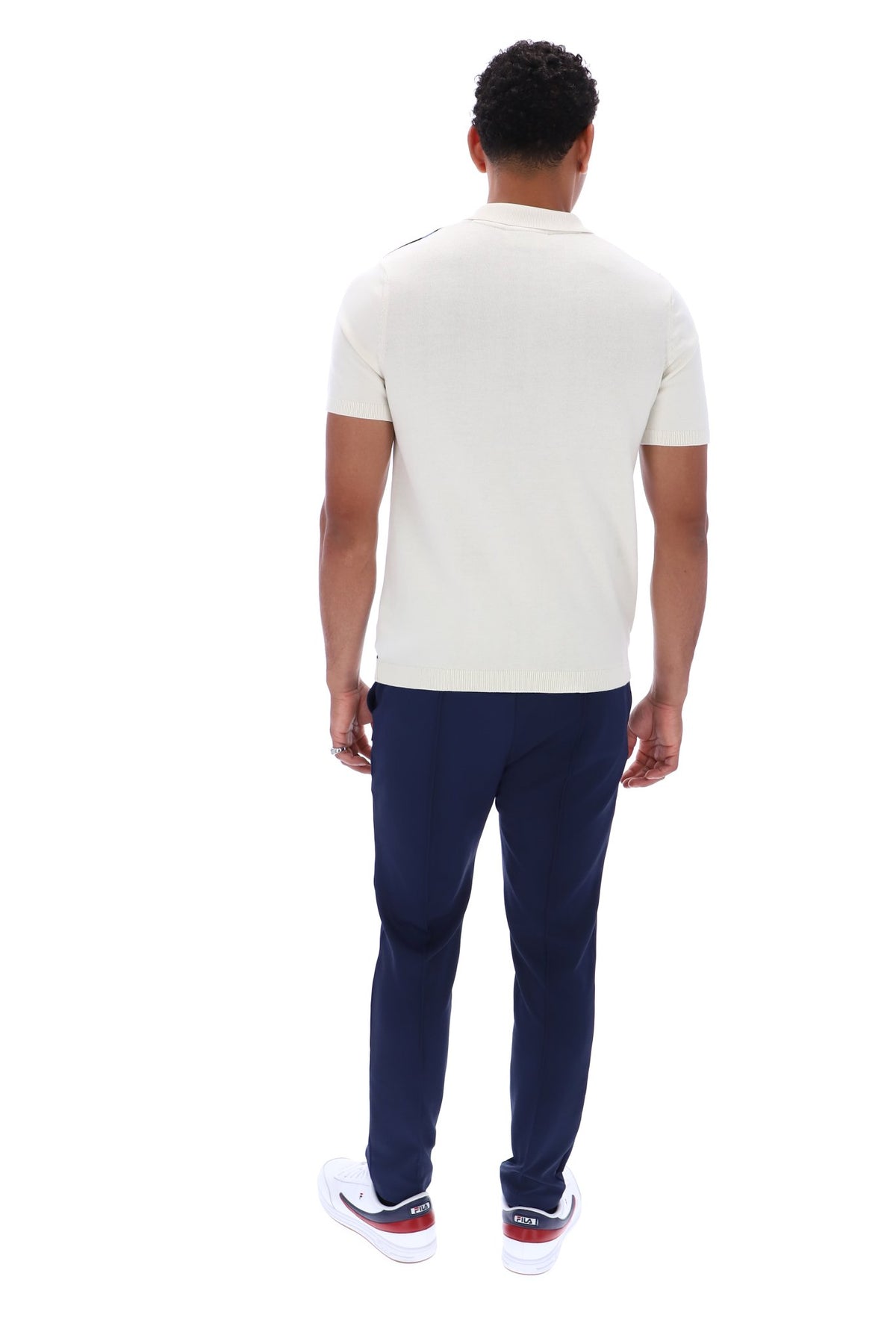 A man viewed from behind wearing a cream short-sleeve polo shirt, navy blue trousers, and white sneakers with red accents, set against a plain white background.