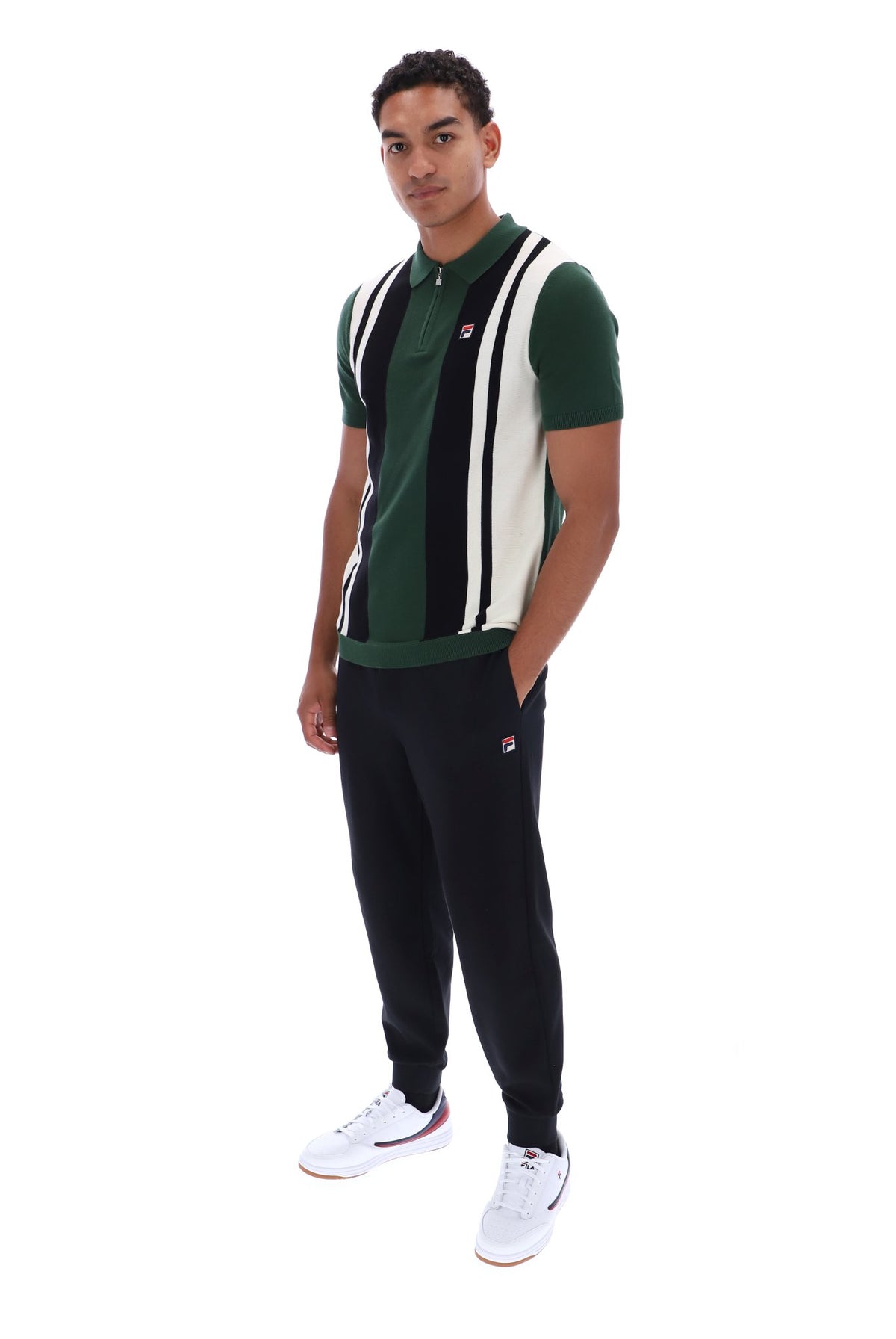 A man modeling a green, black, and white striped polo shirt with matching dark jogger pants and white sneakers, standing against a plain white background.