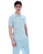 A man wearing a light blue zip-up polo shirt with a FILA gold logo on the chest, on a white background