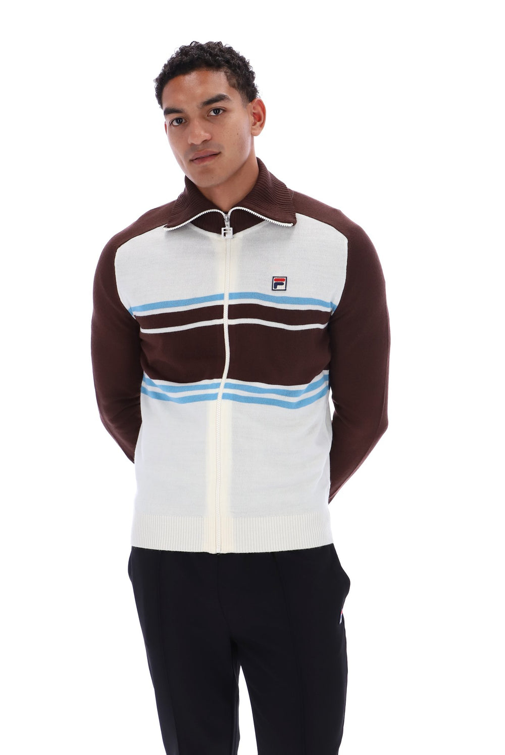 A man wearing a Fila track jacket with a white body, dark brown sleeves and yoke, light blue horizontal stripes, and a white zipper. He is also wearing black pants against a plain white background.