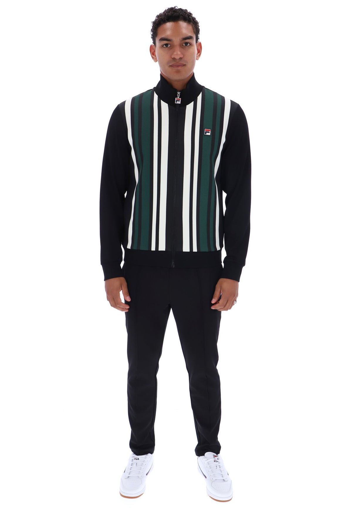 A man modeling a black track jacket with vertical green, white, and black stripes on the front, paired with black sweatpants and white sneakers, against a plain white background.