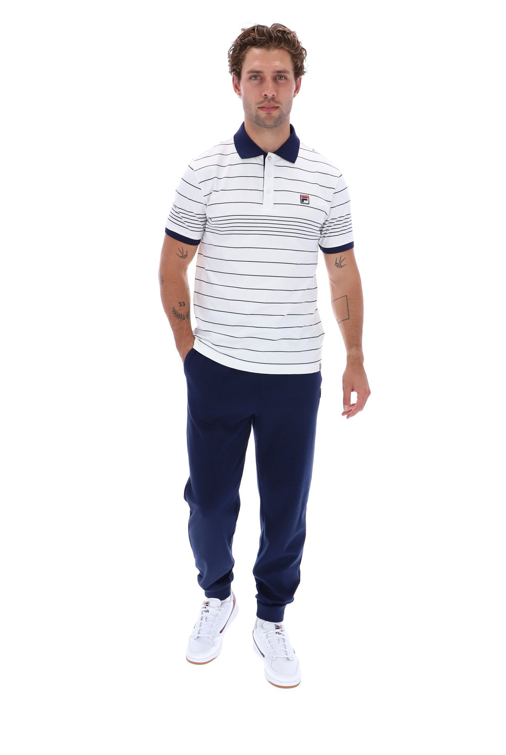 A man modeling a white short-sleeve polo shirt with navy blue horizontal stripes, a navy collar, and a small embroidered logo on the chest, paired with navy jogger pants and white sneakers, against a plain white background.