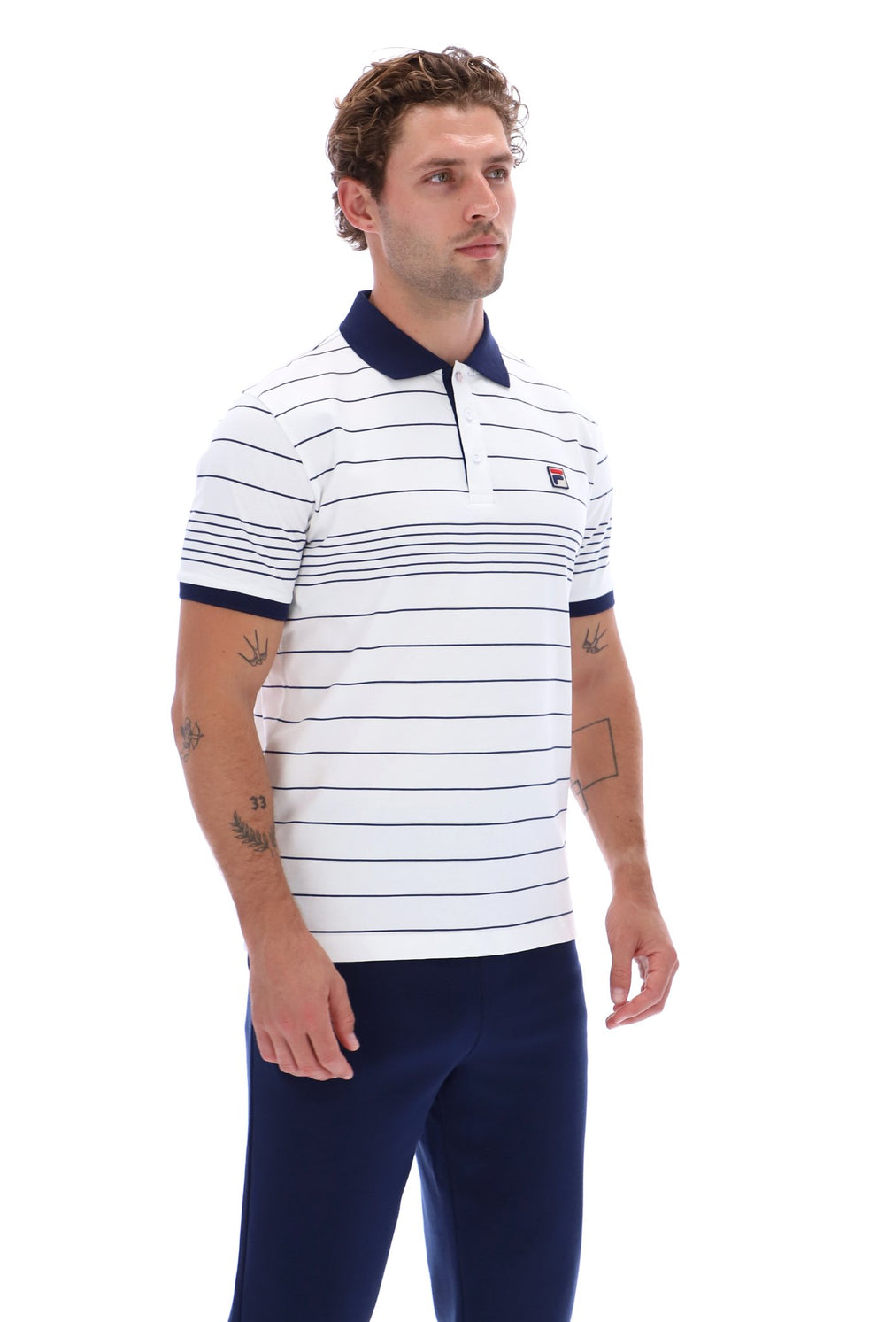 A man wearing a white short-sleeve polo shirt with thin navy blue horizontal stripes, a navy collar, and a small red and white embroidered logo on the chest, paired with navy pants, against a plain white background.