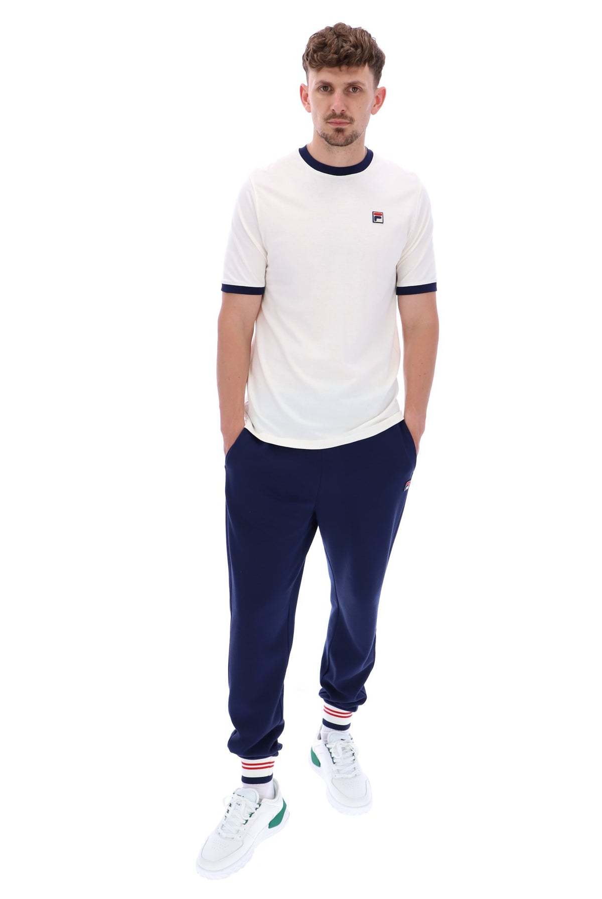 A male model wearing a white and navy ringer t-shirt with the FILA logo on the chest, paired with navy blue joggers on a white background