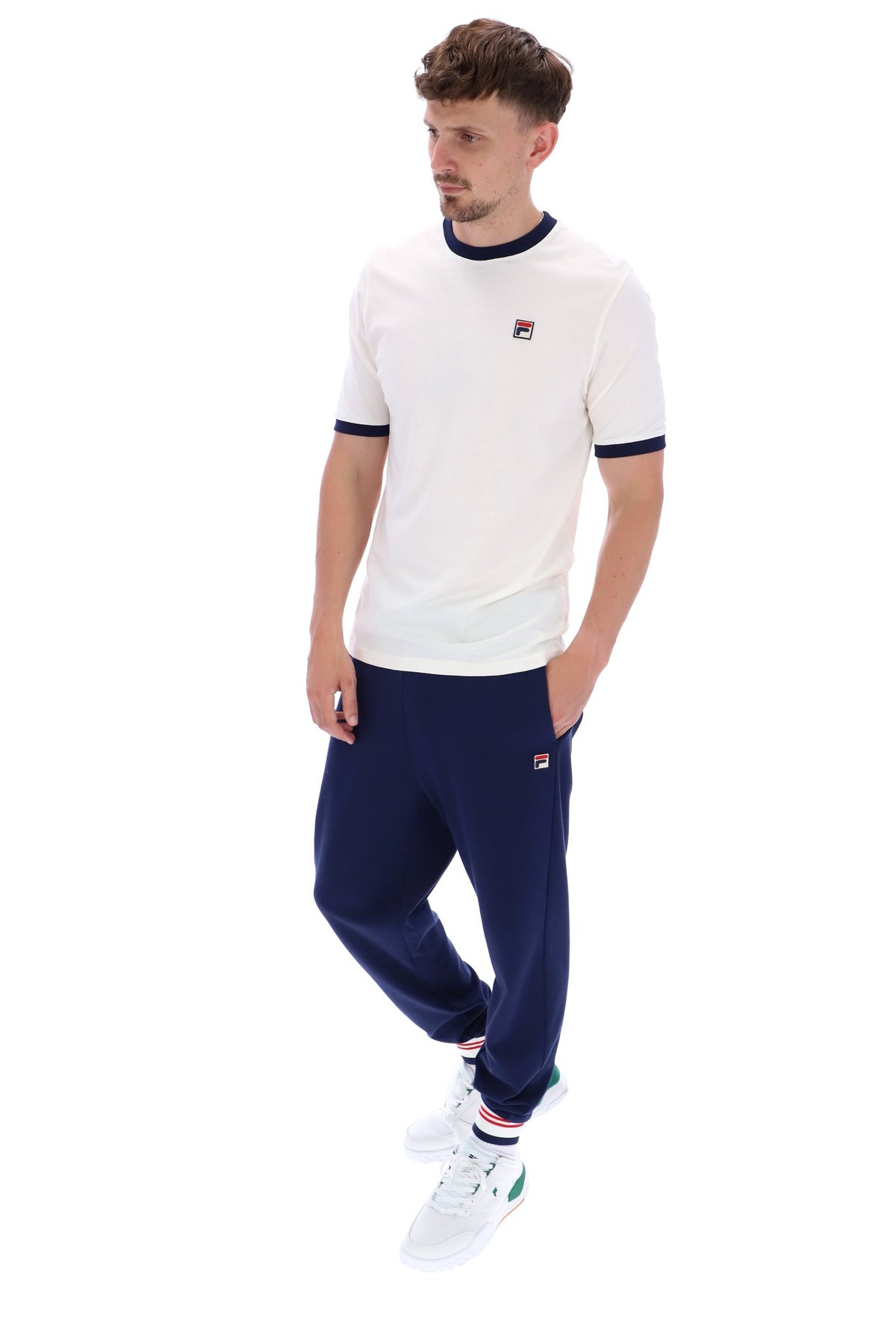 A male model wearing a white and navy ringer t-shirt with the FILA logo on the chest, paired with navy FILA joggers, on a white background
