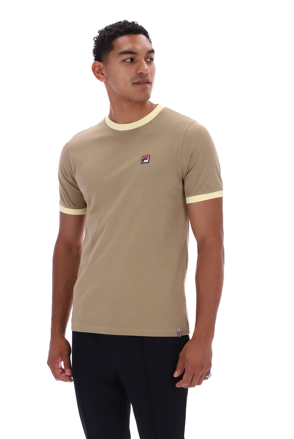 A man wearing a beige ringer t-shirt with cream trim on the collar and sleeve cuffs, featuring a small Fila logo on the left chest, paired with black pants against a white background.