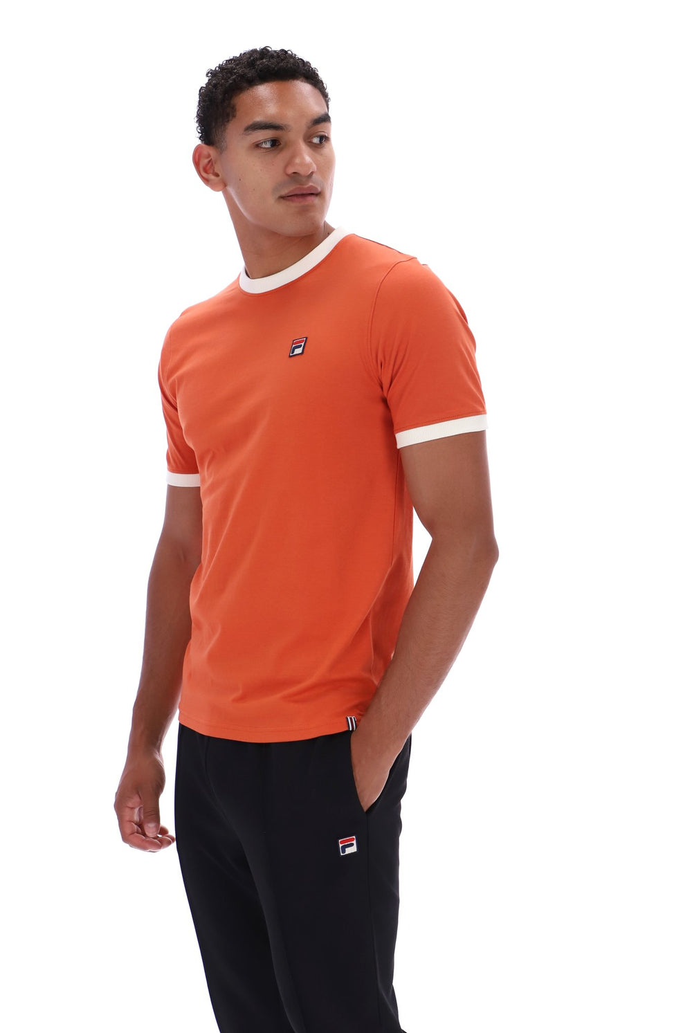 A man wearing a bright orange ringer t-shirt with white trim on the collar and sleeve cuffs, paired with black pants. The shirt features a small embroidered Fila logo on the left chest.