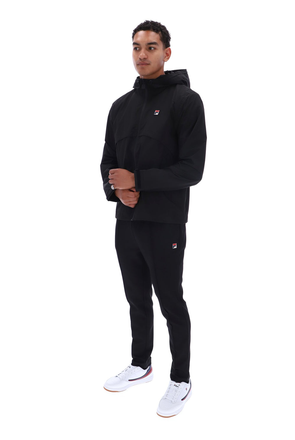 A man modeling a black Fila hooded jacket and matching black track pants with white sneakers, photographed against a solid white background.