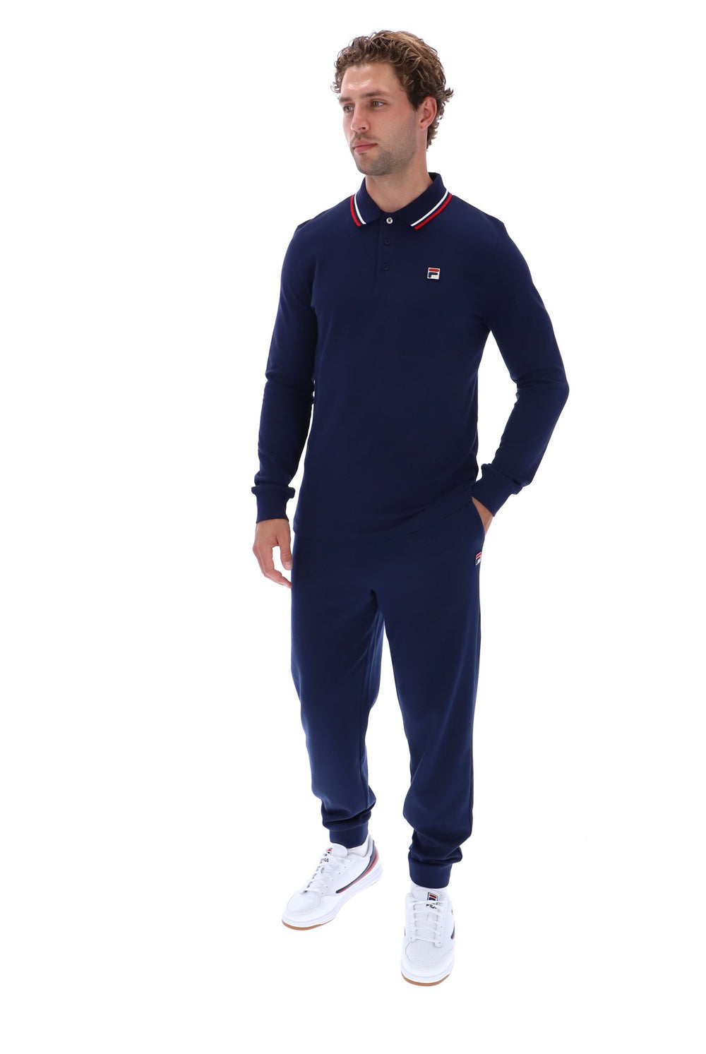 A man modeling a navy blue long-sleeve polo shirt and matching jogger pants set from Fila, featuring a small red and white logo on the chest, paired with white sneakers, against a plain white background.