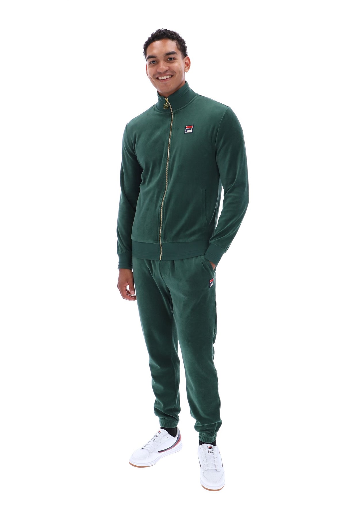 A man modeling a dark green Fila tracksuit, featuring a zip-up jacket with a small red, white, and blue logo on the left chest, and matching jogger pants, against a plain white background.