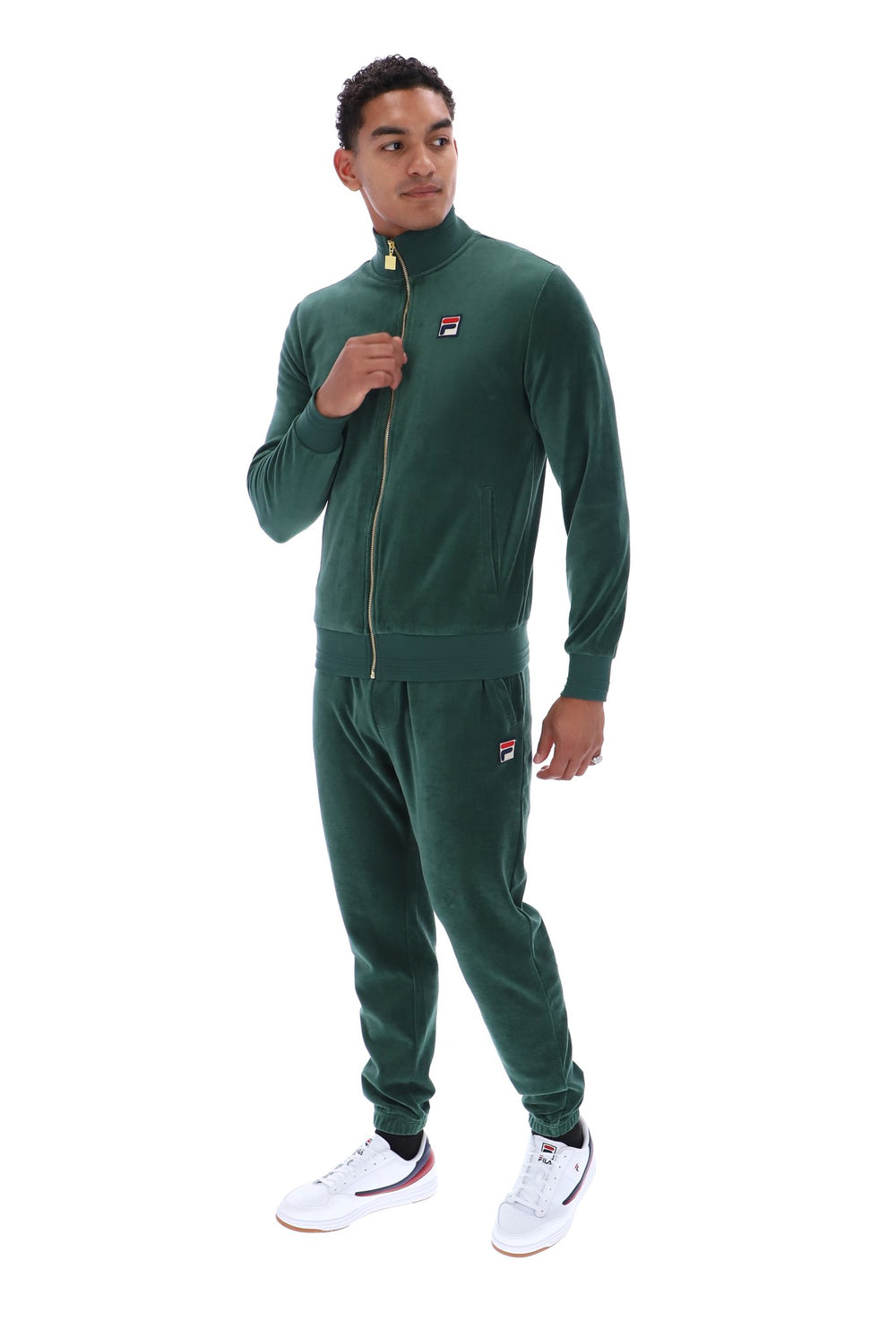A man modeling a dark green Fila tracksuit, featuring a zip-up jacket with a small red, white, and blue logo on the left chest, and matching jogger pants. He is wearing white sneakers with red and blue accents, set against a plain white background.