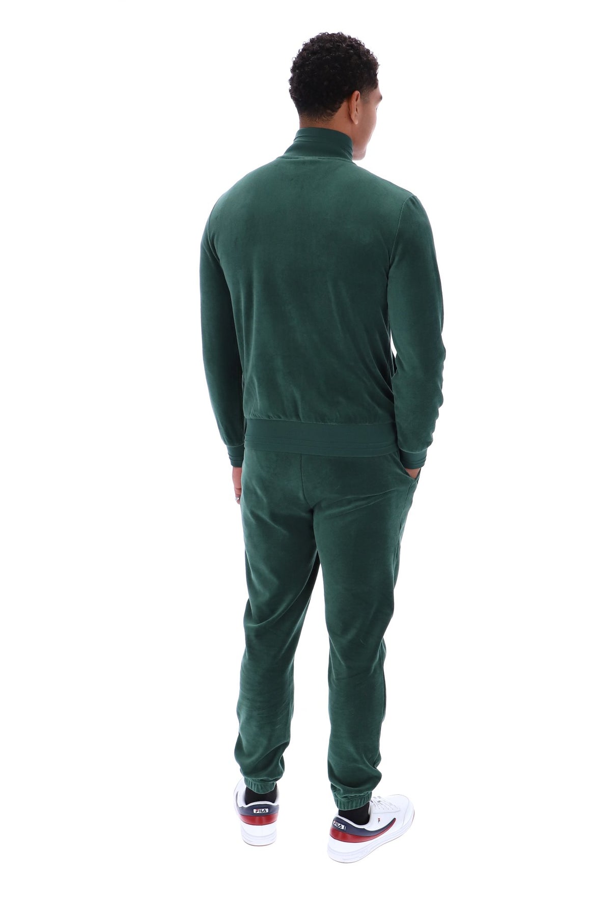 A man viewed from behind wearing a dark green velour tracksuit with a mock neck jacket and matching jogger pants, paired with white sneakers.