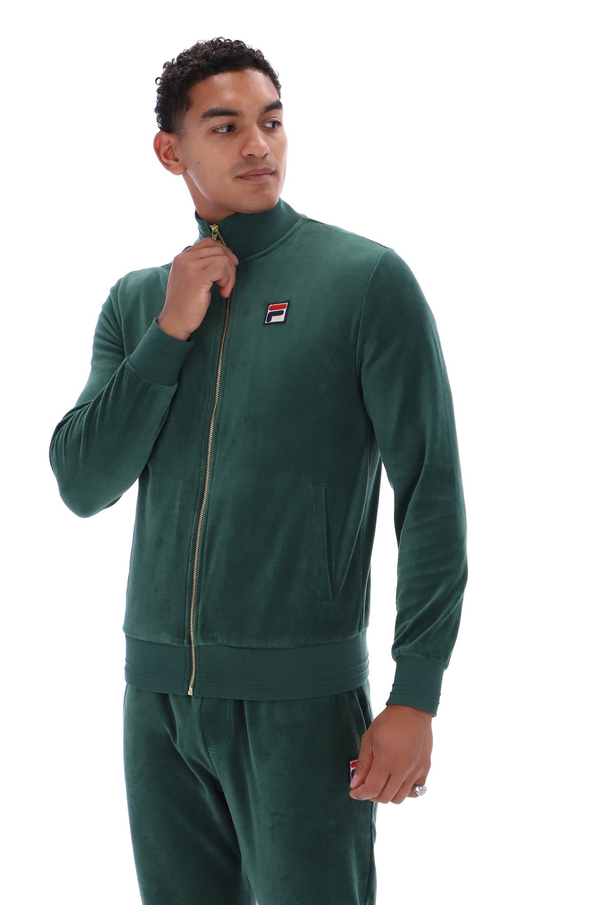 A man wearing a dark green velour tracksuit with a matching zip-up jacket and pants, featuring a small Fila logo patch on the left chest, set against a plain white background.