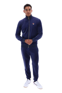 A man modeling a navy blue Fila zip-up track jacket and matching sweatpants set, paired with white sneakers, against a solid white background.