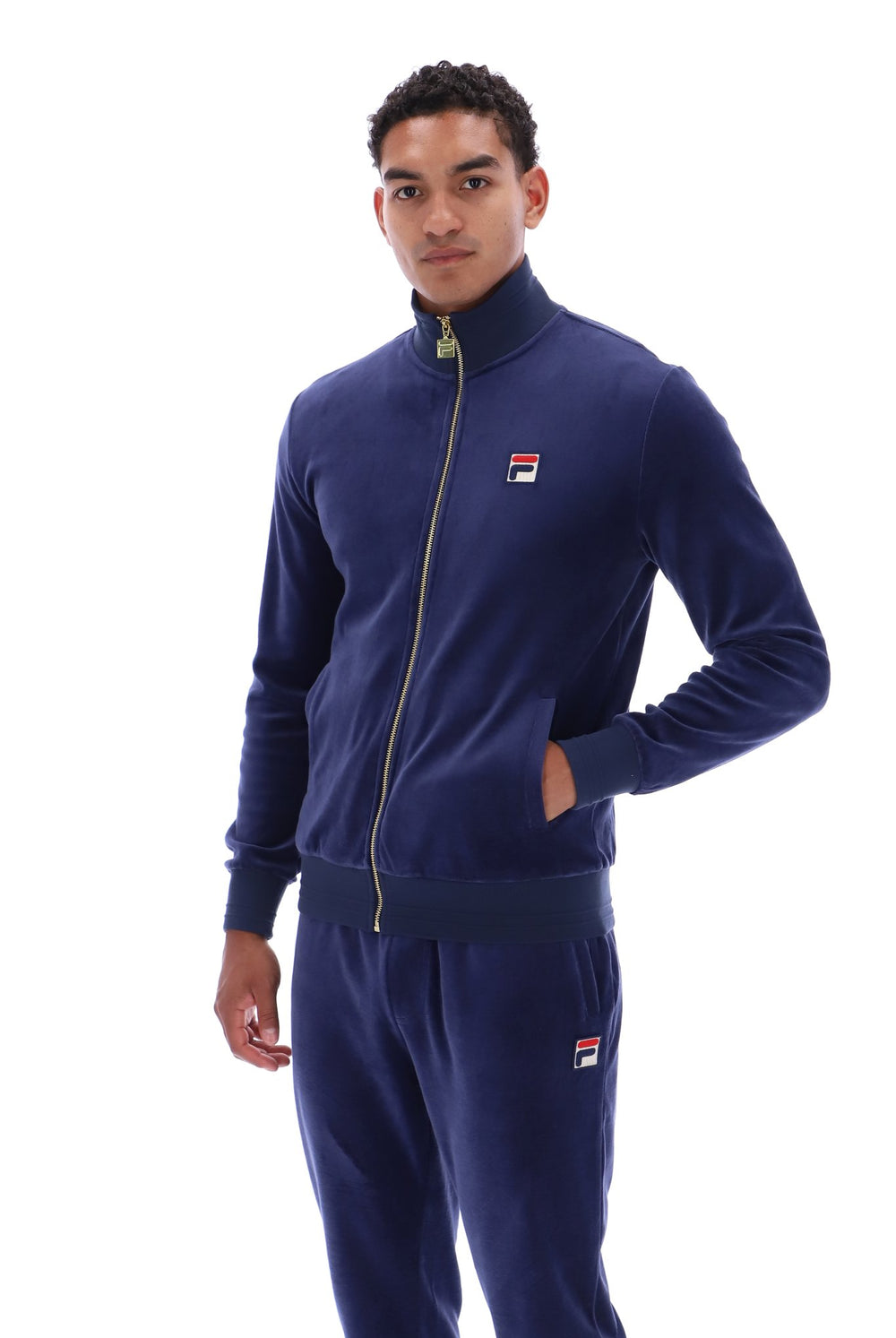 A man wearing a navy blue Fila tracksuit, featuring a zip-up jacket with a small red, white, and blue logo on the left chest, and matching sweatpants with a logo on the lower left leg, shot against a solid white background.