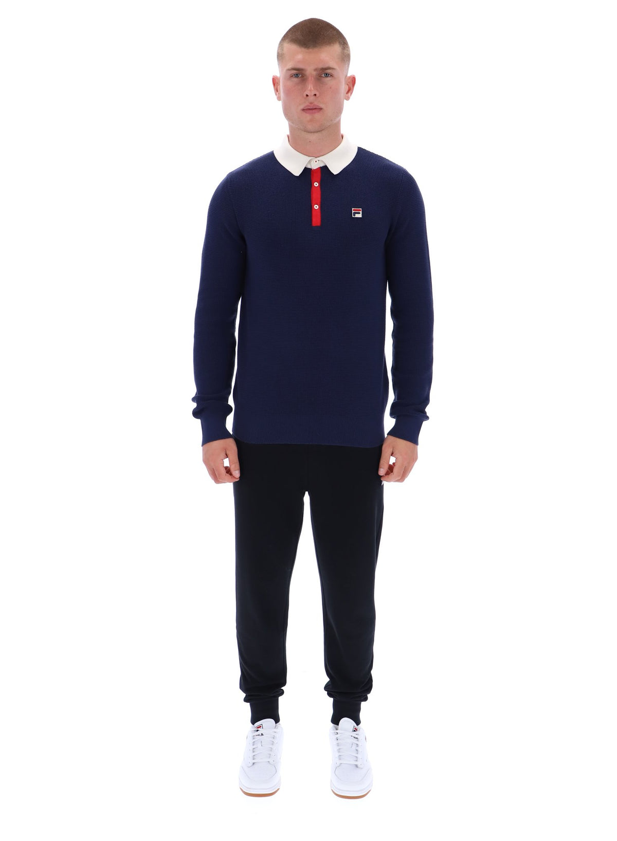 Man wearing a navy sweater with a red and white logo, black pants, and white shoes on a white background