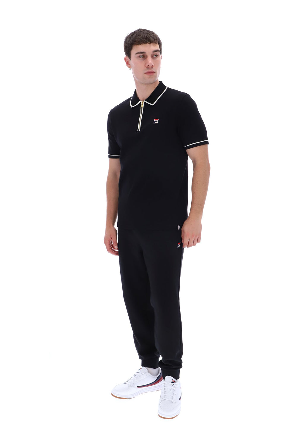 A man modeling a black short-sleeve polo shirt and matching black jogger pants set, featuring white and red trim details, paired with white sneakers, on a solid white background.