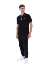 A man modeling a black short-sleeve polo shirt and matching black jogger pants set, featuring white and red trim details, paired with white sneakers, on a solid white background.
