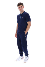 A man modeling a navy blue polo shirt and matching sweatpants set with white sneakers, photographed against a solid white background.