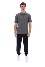 A man modeling a dark gray short-sleeve polo shirt with a half-zip collar, black jogger pants, and white sneakers against a plain white background.