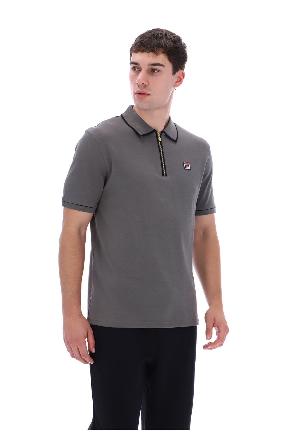 A man wearing a dark gray short-sleeve polo shirt with a black collar, a black zipper at the chest, and a small red and white Fila logo on the left chest, paired with black pants against a white background.