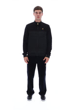 A man modeling a black zip-up jacket and matching black sweatpants against a solid white background.