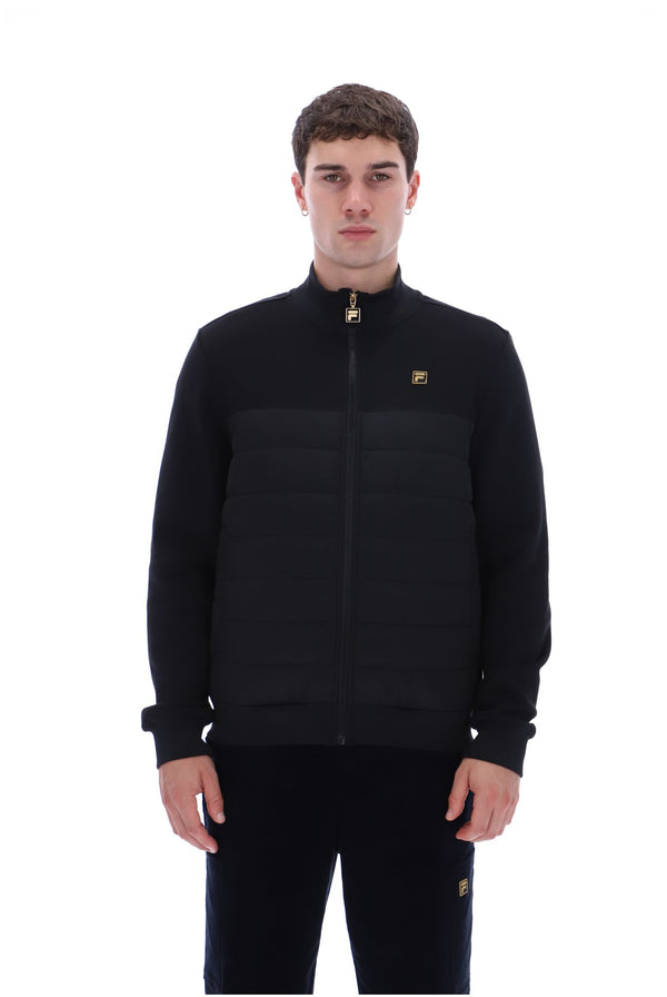 A man wearing a black zip-up jacket with horizontal quilting on the lower half and a small gold logo on the left chest, paired with black pants, against a plain white background.