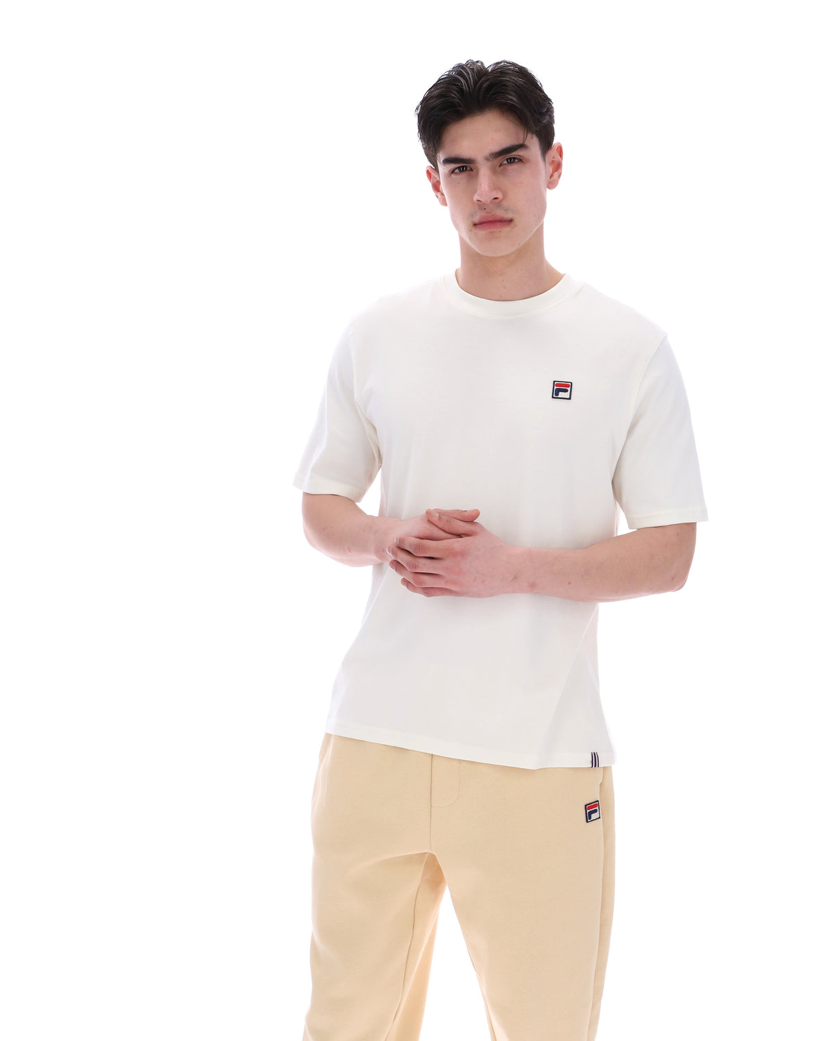 Person wearing a white t-shirt with a small logo and beige pants on a white background