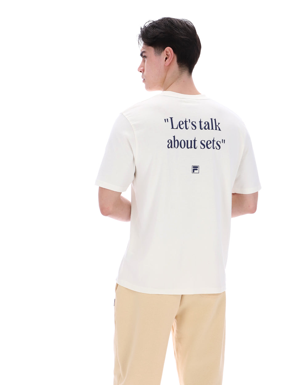 Person wearing a white t-shirt with text reading "Let's talk about sets" on the back, with a small black FILA logo standing against a white background